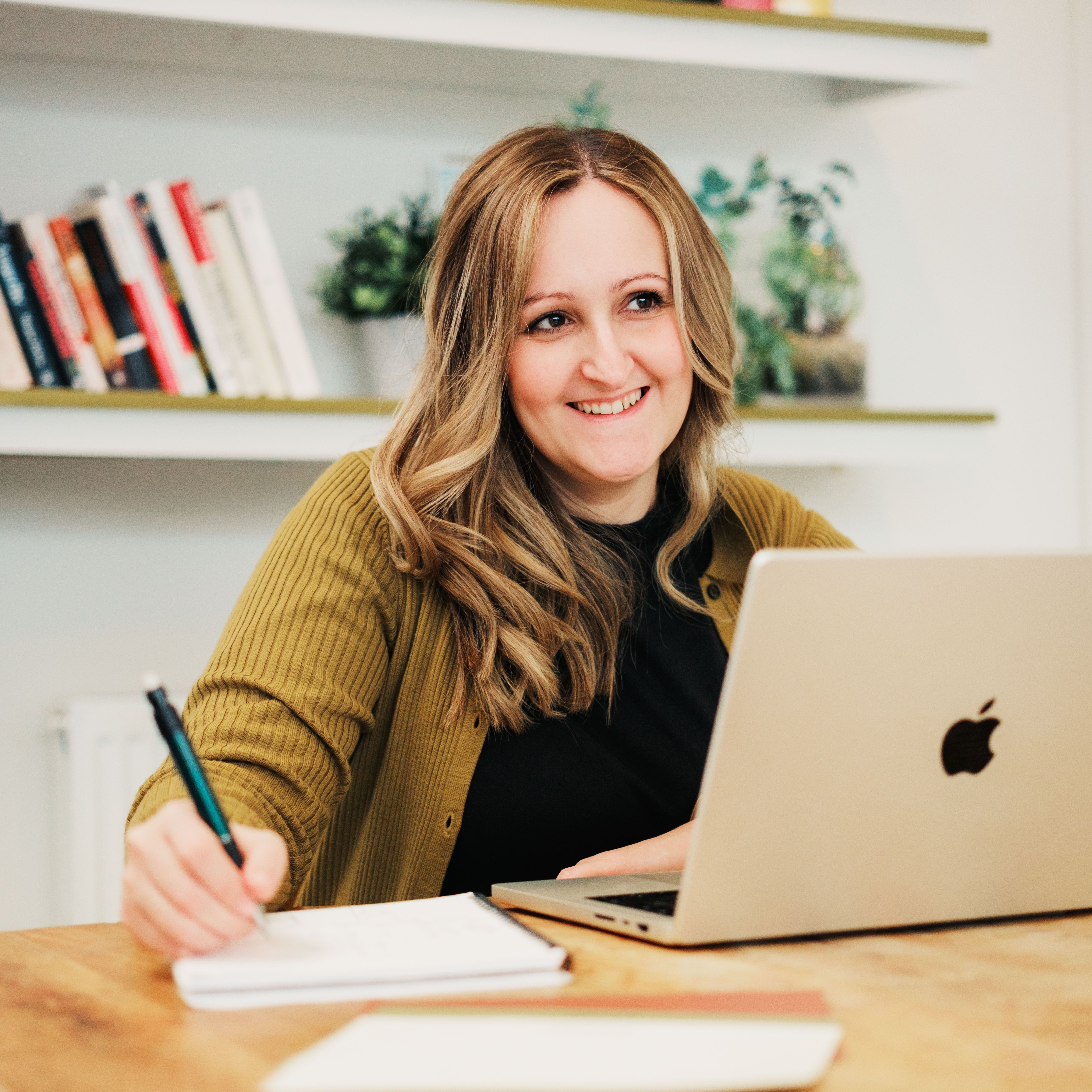 Becca sat at a wood topped desk with a pencil, notebook and laptop. Becca is a white female with wavy brown hair with blonde highlights. She is wearing a mustard cardigan over a black top. She is smiling. Books and plants are on the shelves in the background.