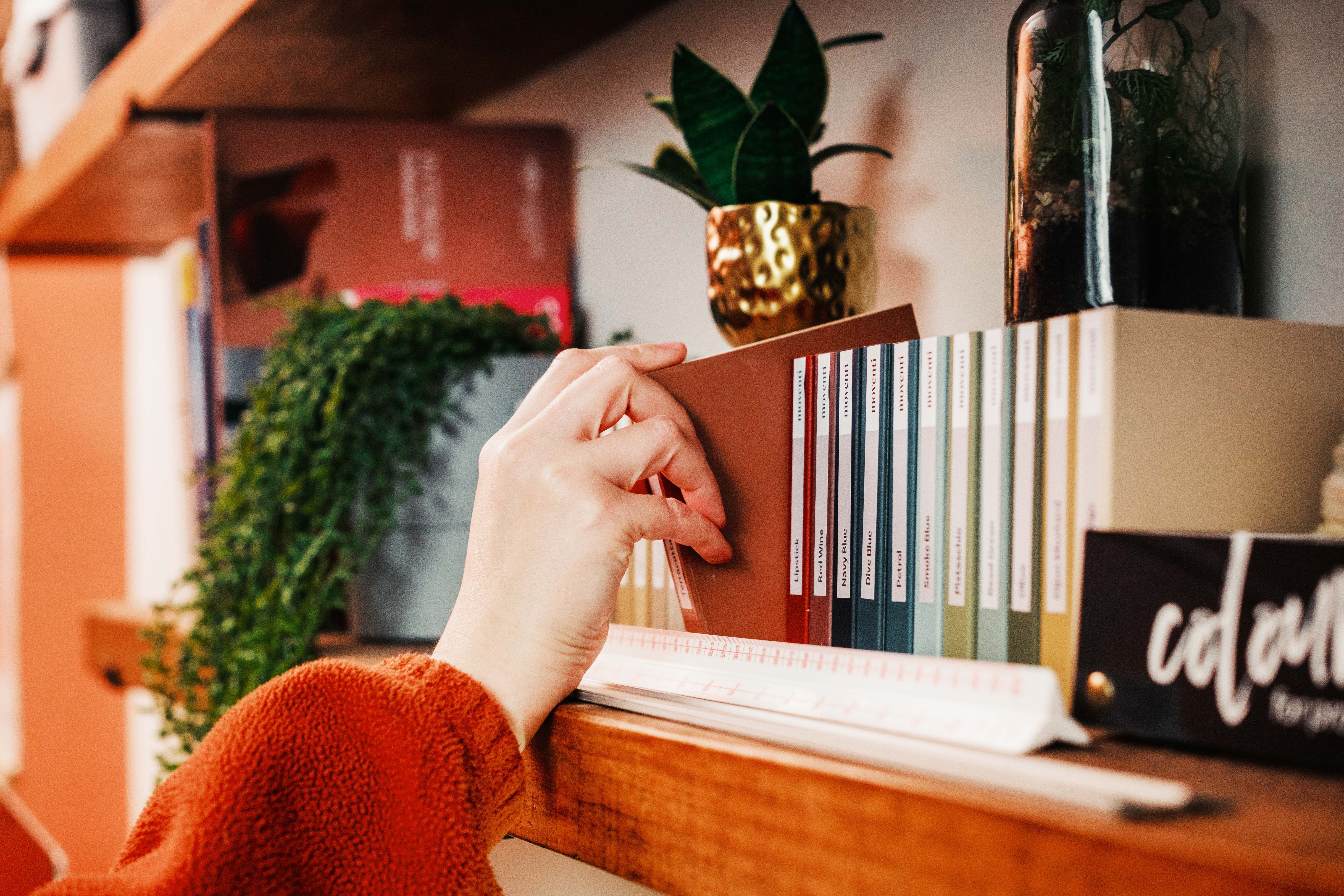 Hand taking an MFC sample from a set of samples on a wooden shelf. A scale ruler, small plants, books, and colour swatches are also in frame.